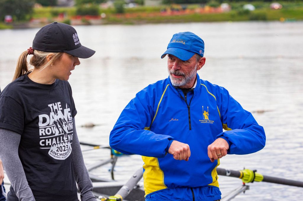 A man and a woman standing in front of a lake. The man is making a motion with his arms as if demonstrating rowing.